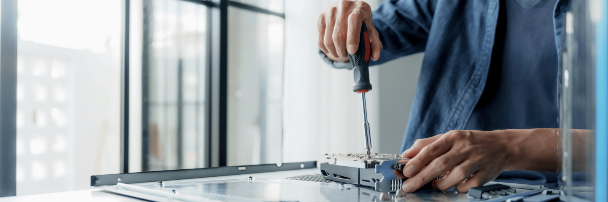 A close-up of a person's hands using a screwdriver on an electronic component on a workbench, with a blurred office background.